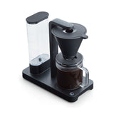 A PERFORMANCE BLACK coffee maker with a transparent water reservoir and a glass carafe filled with coffee. Featuring unique technology for the perfect temperature, it boasts two award badges: one with a laurel wreath indicating "Best i Test Tek.no" and another that reads "Testvinner" with stars and a ribbon symbol.